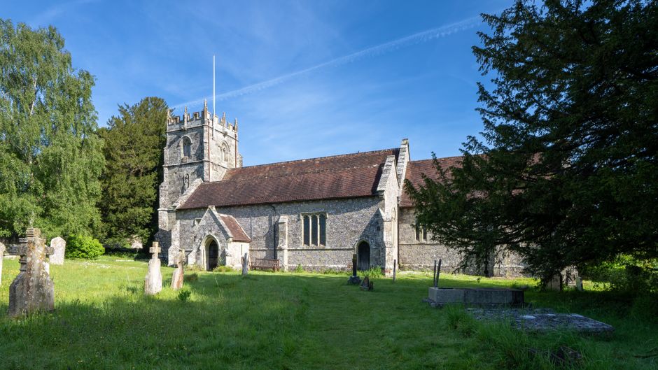 A historic stone church with a red tiled roof and a prominent square tower topped with battlements, situated amid lush green grass and trees on a bright, sunny day. The church features narrow, arched 